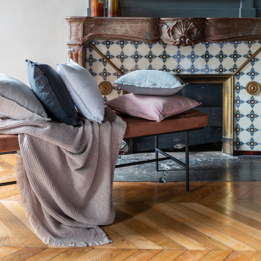 five pillows of various colours on a brown bench in front of a fireplace